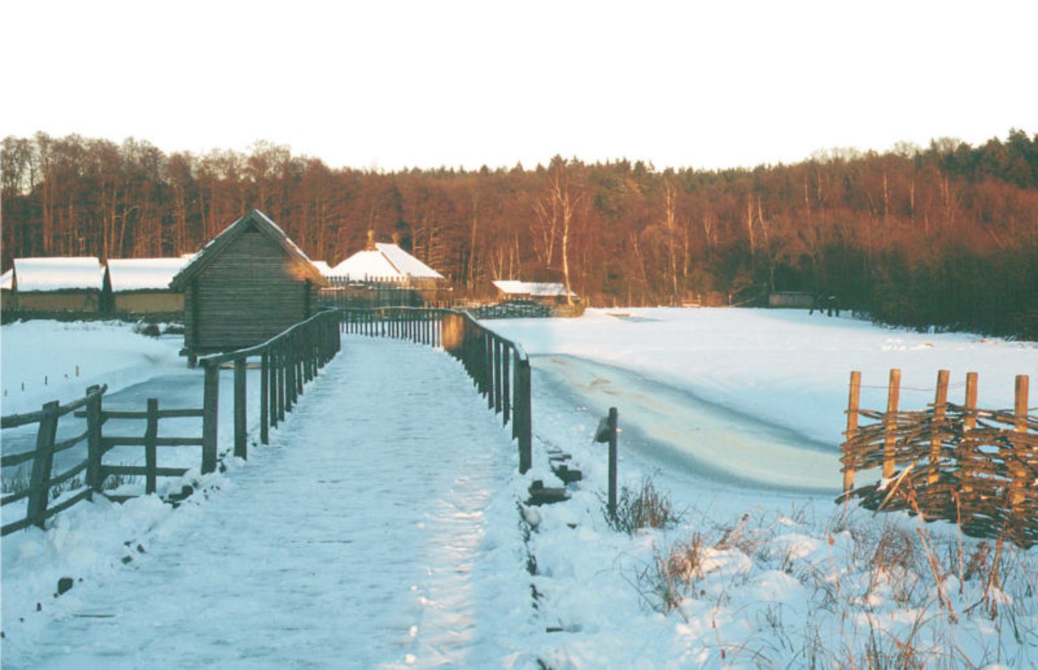 Das Freigelände im Winter. (© LAKD M-V/LA) Die Landschaft ist von Schnee bedeckt. Hinter einer hölzernen Brücke, an der sich ein kleines Haus mit Holzwänden und Strohdach befindet, sind weitere Häuser mit schneebedeckten Dächern zu sehen. (© LAKD M-V/LA)