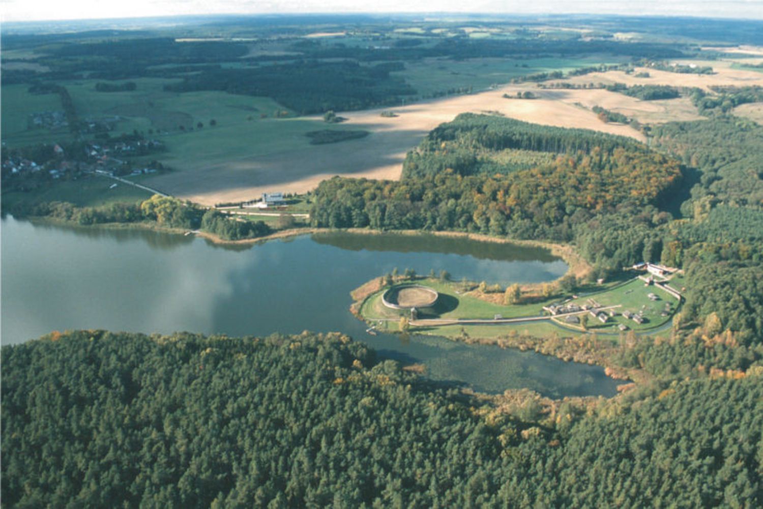 Das Freilichtmuseum aus der Vogelperspektive, Zustand um 2005. (© LAKD M-V/LA) Aus der Vogelperspektive ist eine Landschaft mit Feldern, Wäldern und einem See zu sehen. Auf einer Halbinsel im See liegt ein Burgwall, im Hintergrund links das Dorf Groß Raden. (© LAKD M-V/LA)