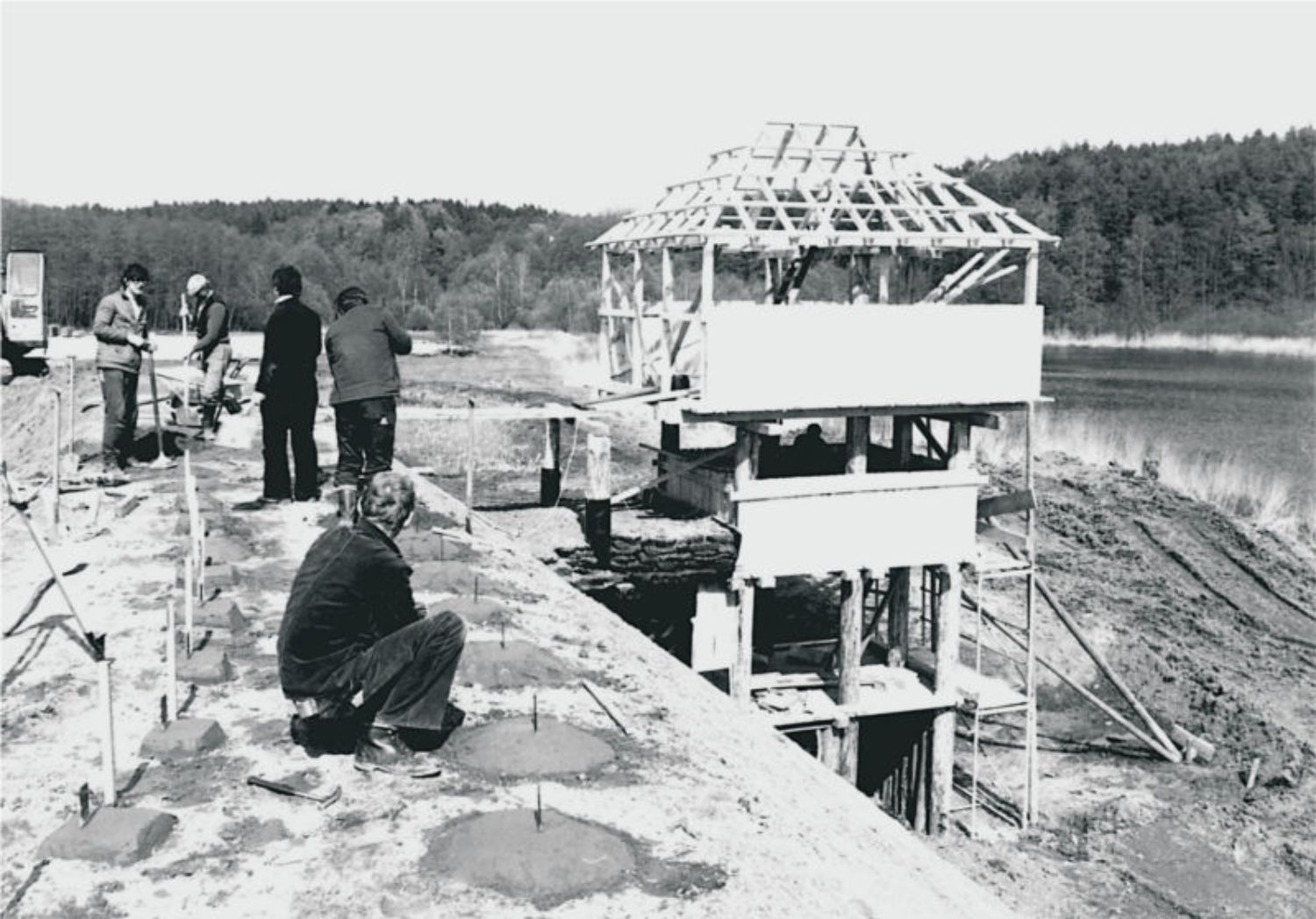 Der hölzerne Treppenturm am Burgwall. Für ihn gab es kein originales Vorbild. (© LAKD M-V/LA) Auf der Krone eines im Bogen verlaufenden Walles stehen und knien Menschen. Außen vor dem Wall steht ein hölzerner Turm mit mehreren Etagen und einem Walmdach, von dem eine Verbindung zum Wall führt. (© LAKD M-V/LA)