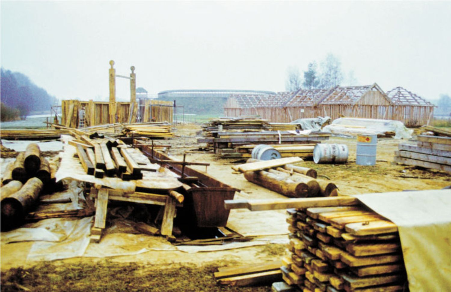 Das Freigelände im Bau. Im Hintergrund Tempel, Burgwall und die ersten Flechtwandhäuser. (© LAKD M-V/LA) Im Vordergrund liegen Holzstapel, Fässer und anderes Baumaterial. Im Hintergrund stehen die hölzernen Gerüste mehrerer Häuser. (© LAKD M-V/LA)