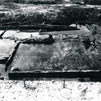 Der Blick fällt von schräg oben auf eine Grabungsfläche, die teilweise unter Wasser steht. In der Grabungsfläche liegen mehrere Hölzer, außerdem sind die unteren Teile geflochtener Wände zu sehen. Eine größere Fläche ist mit dicht gepackten Zweigen bedeckt. (© LAKD M-V/LA )